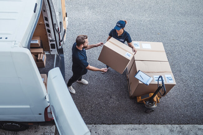 Two people are loading parcels into an open delivery van. The parcels are stacked on a hand pallet truck in front of the open delivery van.